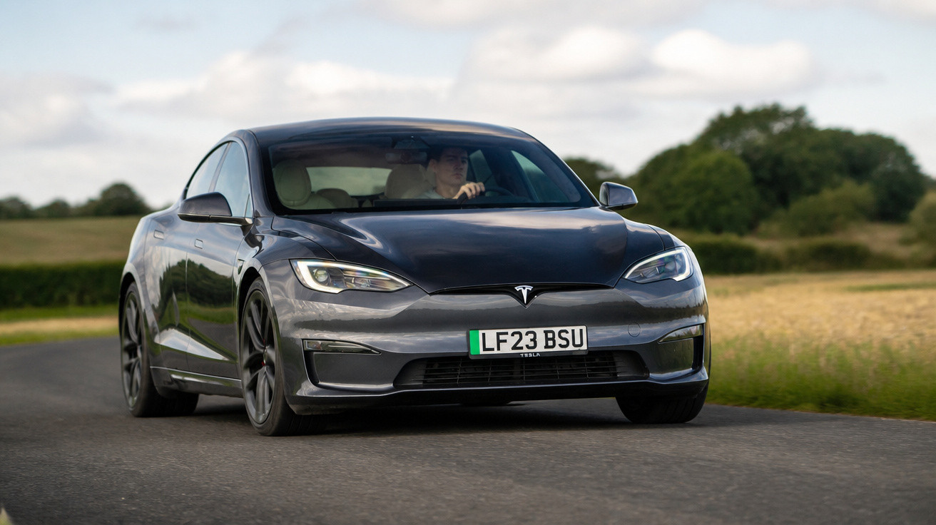 Carro Tesla cinza escuro trafegando em estrada rural com motorista visível ao volante.