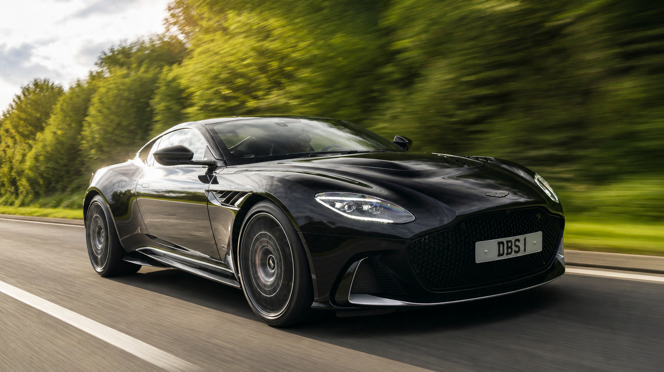 Carro esportivo preto Aston Martin DBS em alta velocidade na estrada com árvores ao fundo.