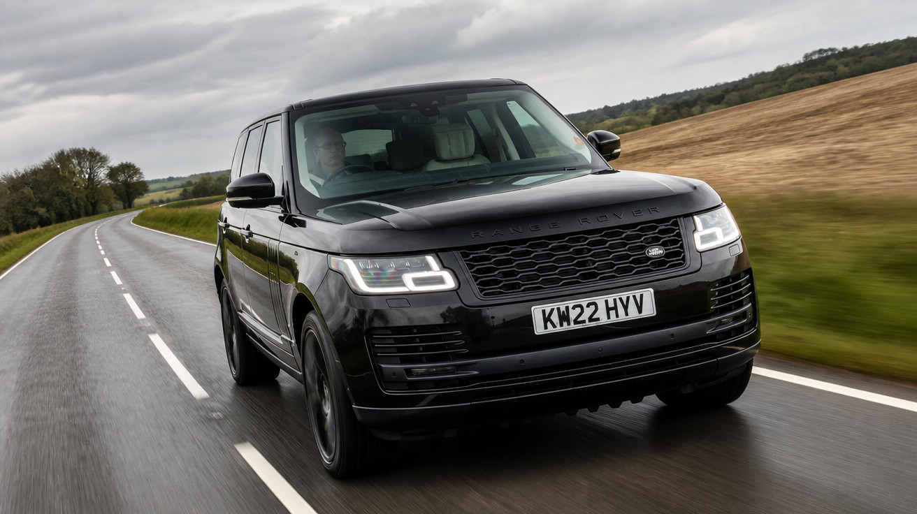 SUV preto Range Rover rodando em estrada molhada com céu nublado e campos ao redor.