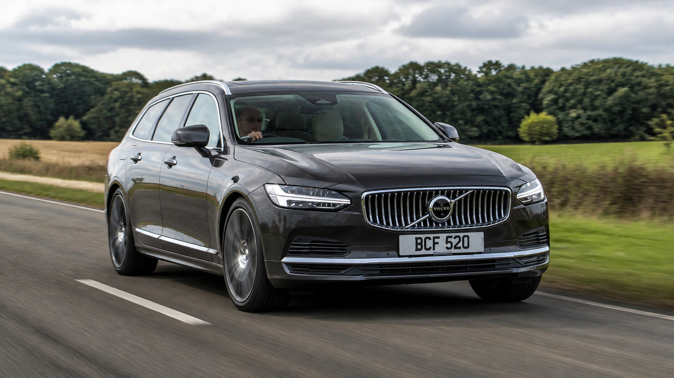 Carro Volvo preto modelo perua em estrada asfaltada com vegetação ao fundo em dia nublado.