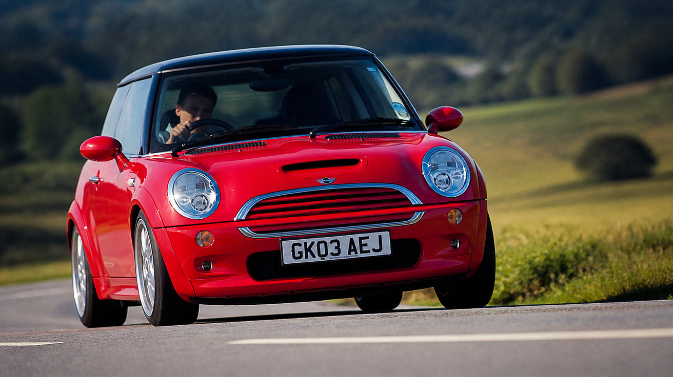 Carro vermelho Mini Cooper com motorista, em estrada asfaltada com paisagem verde ao fundo.