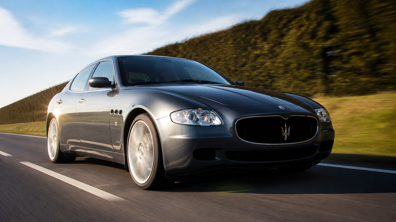 Carro esportivo cinza escuro Maserati em movimento na estrada com vegetação ao fundo em dia ensolarado.
