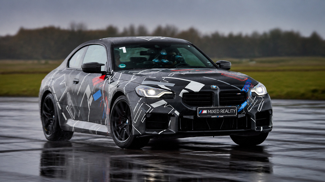 Carro esportivo BMW M com pintura camuflada em pista molhada sob céu nublado.