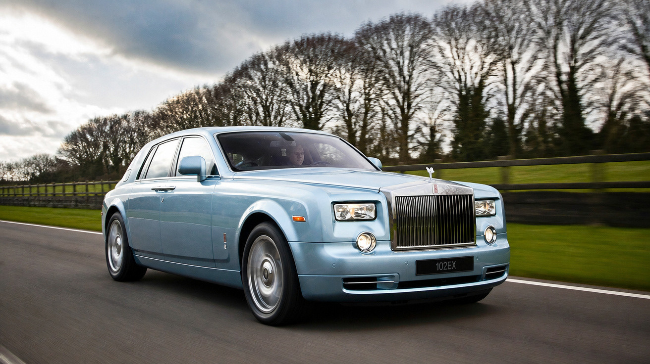 Carro de luxo Rolls-Royce azul claro em movimento em estrada com árvores ao fundo em dia nublado.