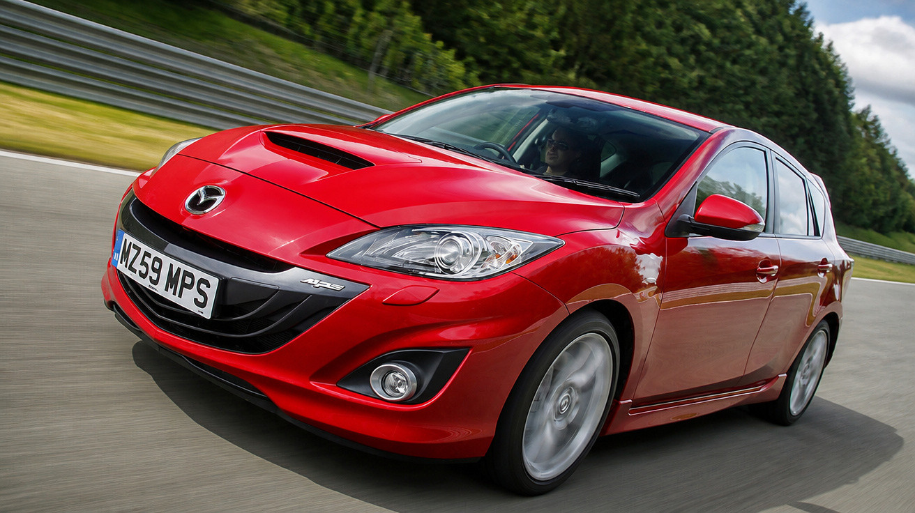 Carro vermelho Mazda hatchback em alta velocidade em pista de corrida com árvores ao fundo.