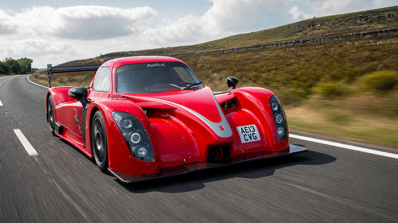 Carro esportivo vermelho com aerofólio grande trafegando em estrada asfaltada com colinas ao fundo.