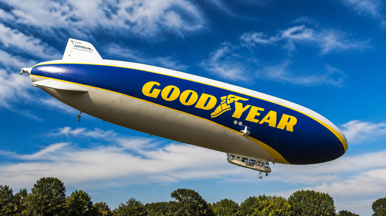 Dirigível Goodyear voando sobre árvores com céu azul e nuvens ao fundo.