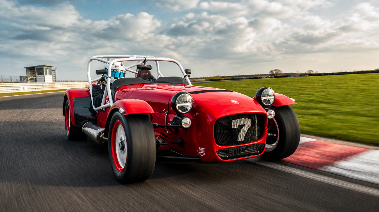 Carro esportivo vermelho aberto com número 7 em pista de corrida com piloto usando capacete branco.