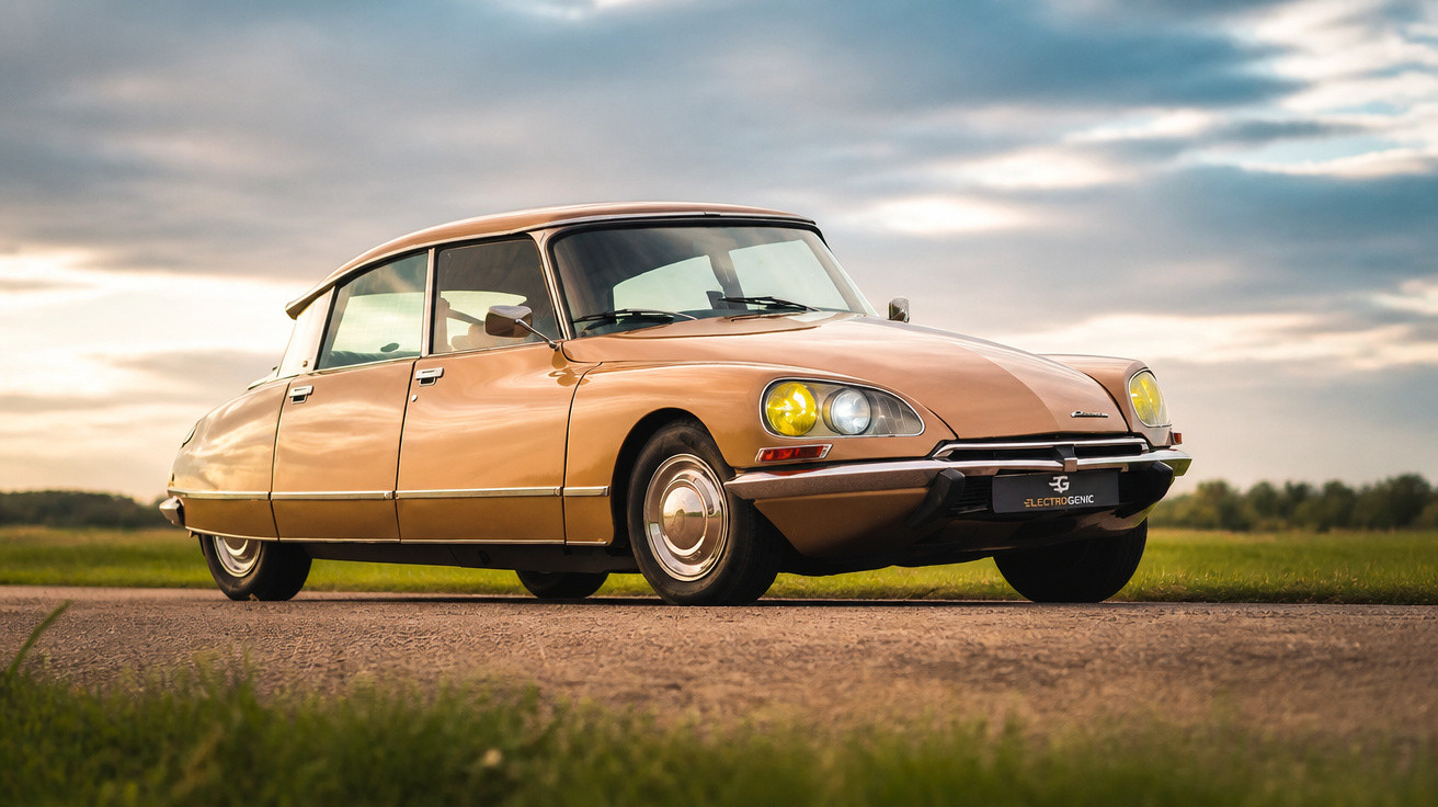 Carro clássico Citroën DS bege estacionado em estrada com céu nublado ao fundo, em ângulo baixo.