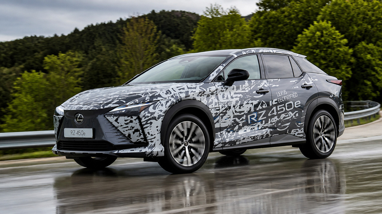 Carro elétrico Lexus RZ 450e camuflado em teste, em estrada molhada com vegetação ao fundo.