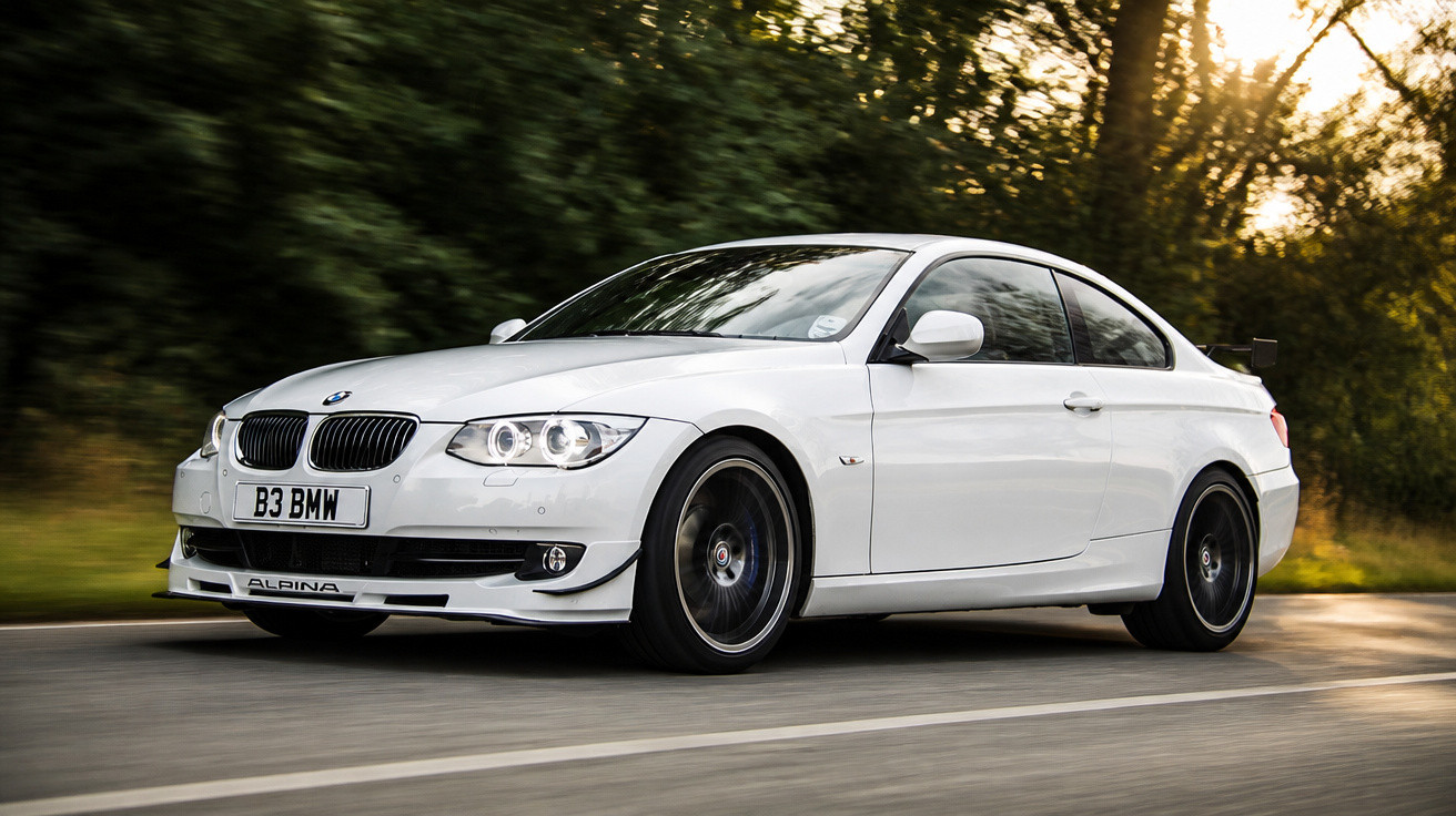 Carro BMW branco modelo Alpina em alta velocidade na estrada com fundo de árvores ao entardecer.