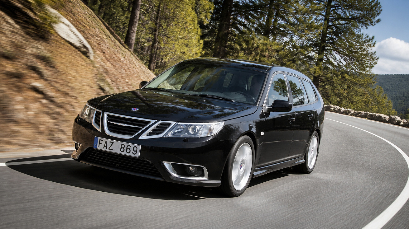 Carro preto Saab modelo station wagon dirigindo em estrada sinuosa cercada de árvores e pedras.