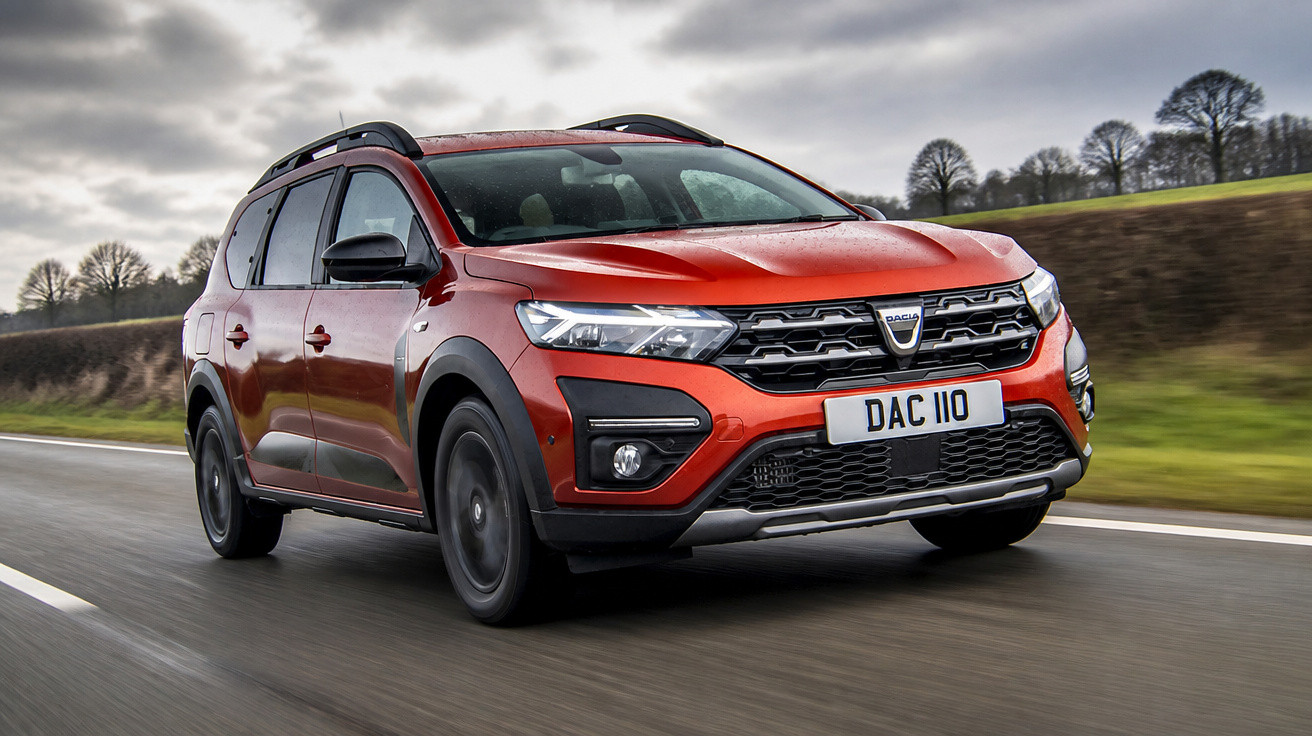 Carro SUV Dacia vermelho em movimento em estrada com céu nublado e árvores ao fundo.