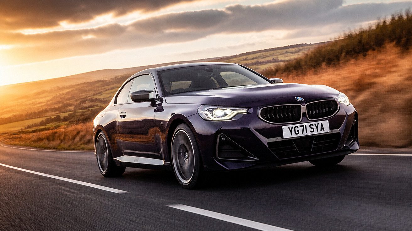 Carro esportivo BMW preto dirigindo em estrada ao pôr do sol com colinas ao fundo.