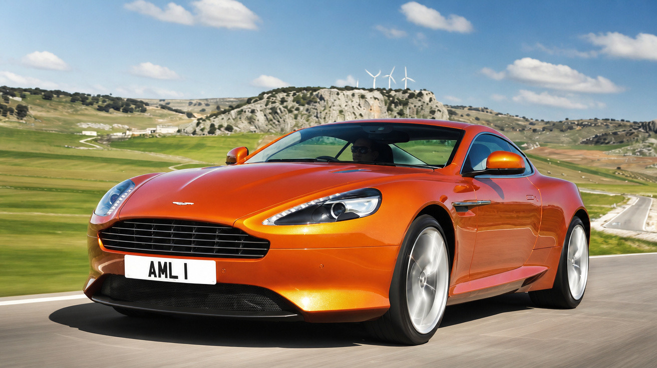 Carro esportivo laranja Aston Martin em estrada com paisagem montanhosa ao fundo.