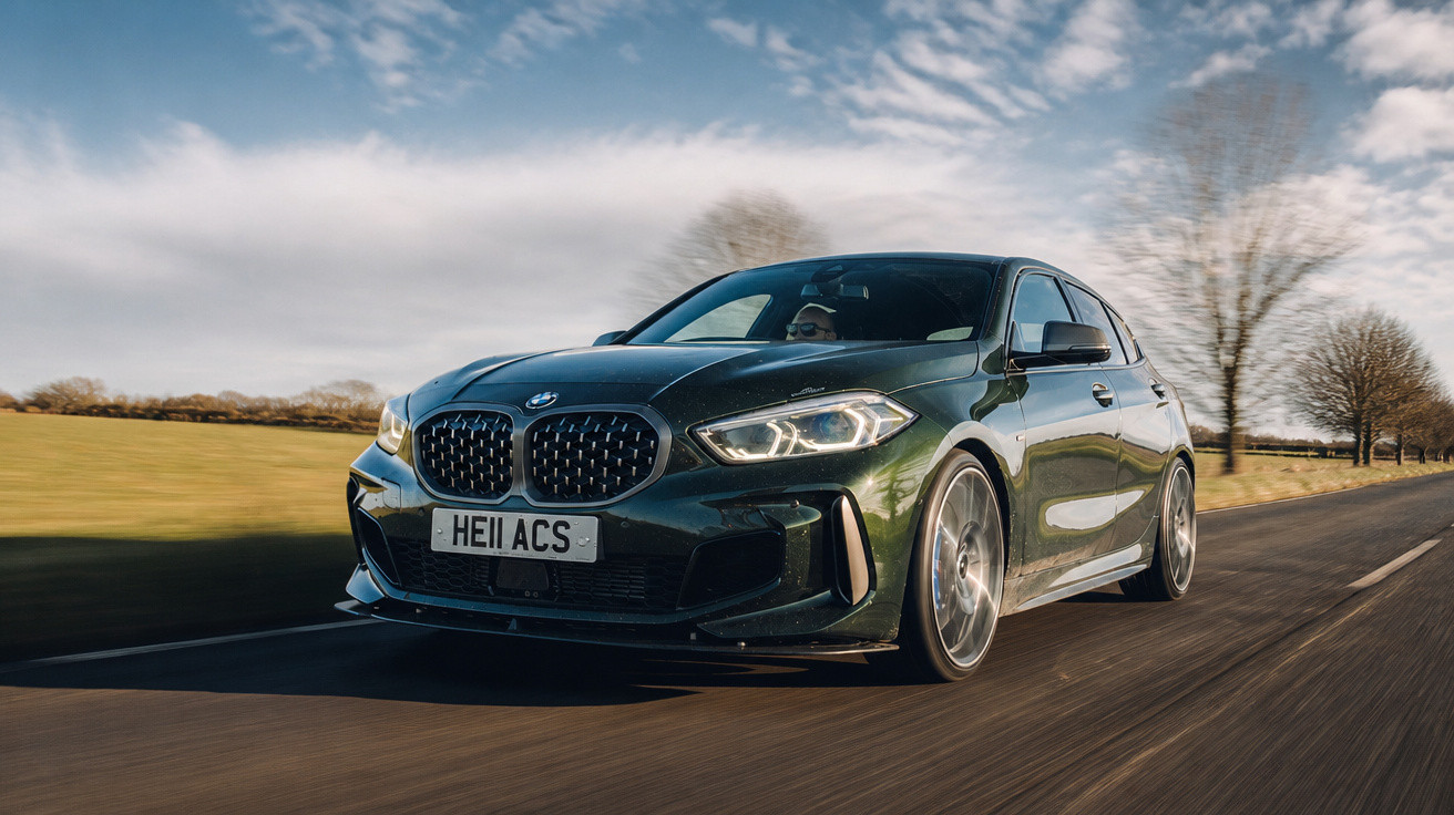 Carro BMW preto em movimento em estrada rural com céu parcialmente nublado e árvores ao fundo.