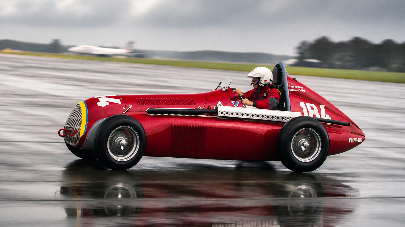 Carro de corrida antigo vermelho com piloto de capacete branco em pista molhada, em movimento.