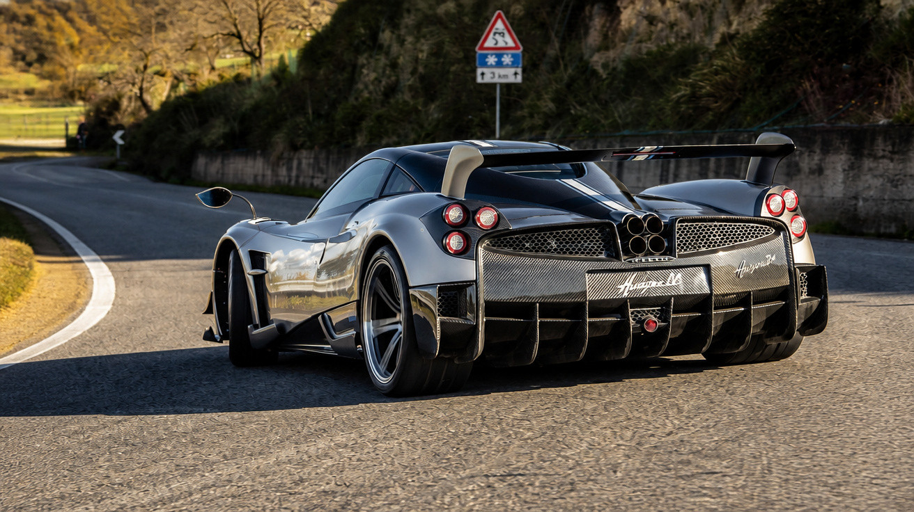 Carro esportivo preto Pagani Huayra em uma estrada curvada com sinal de perigo à frente.