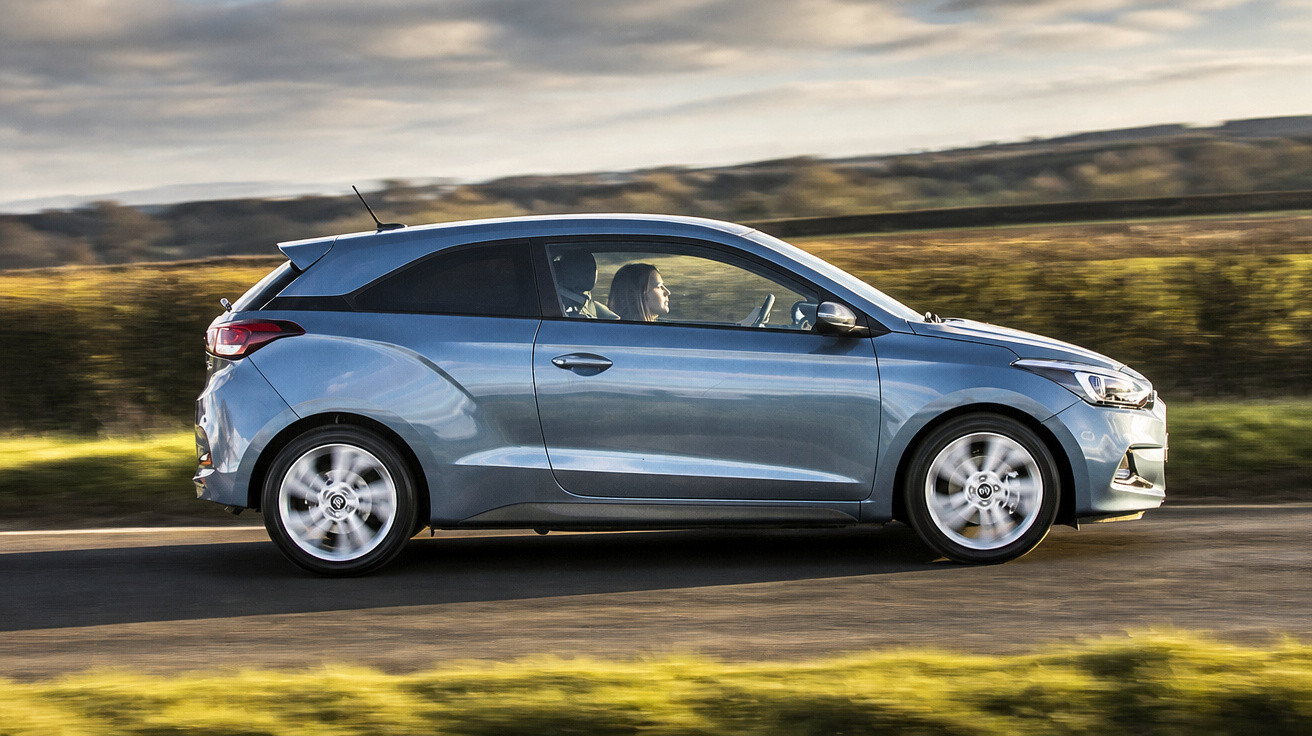 Carro hatchback cinza em movimento em estrada rural com pessoas dentro e paisagem ao fundo.