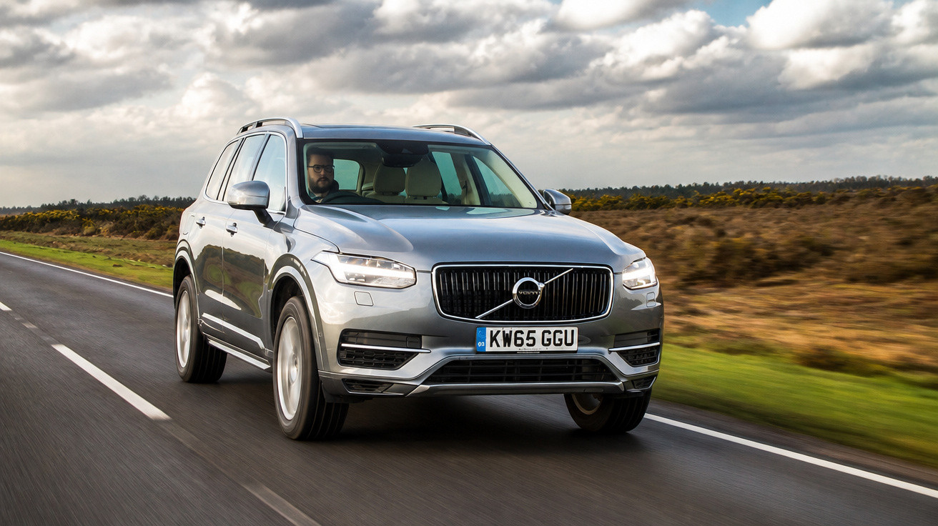 Carro SUV cinza Volvo em movimento em estrada asfaltada cercada por vegetação e céu nublado.