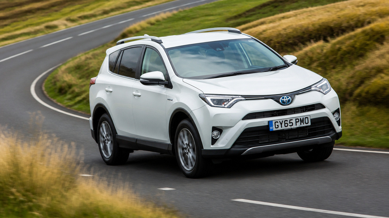 Carro SUV branco Toyota RAV4 em curva de estrada asfaltada cercada por grama verde e seca.