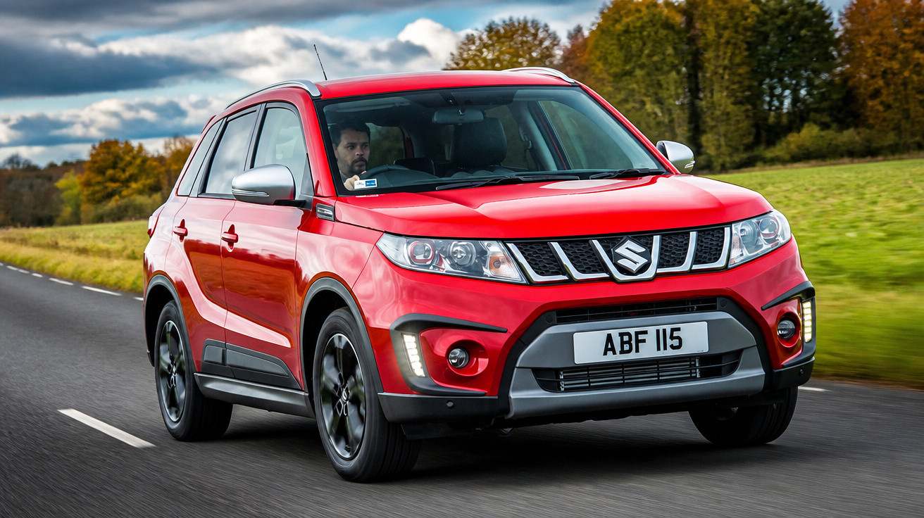 Carro SUV vermelho Suzuki Vitara trafegando em estrada cercada por áreas verdes e céu nublado.
