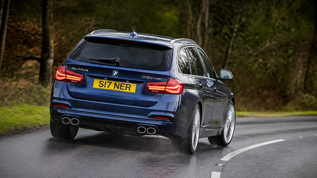 Carro BMW Alpina azul escuro dirigindo em estrada molhada com árvores ao redor à tarde.
