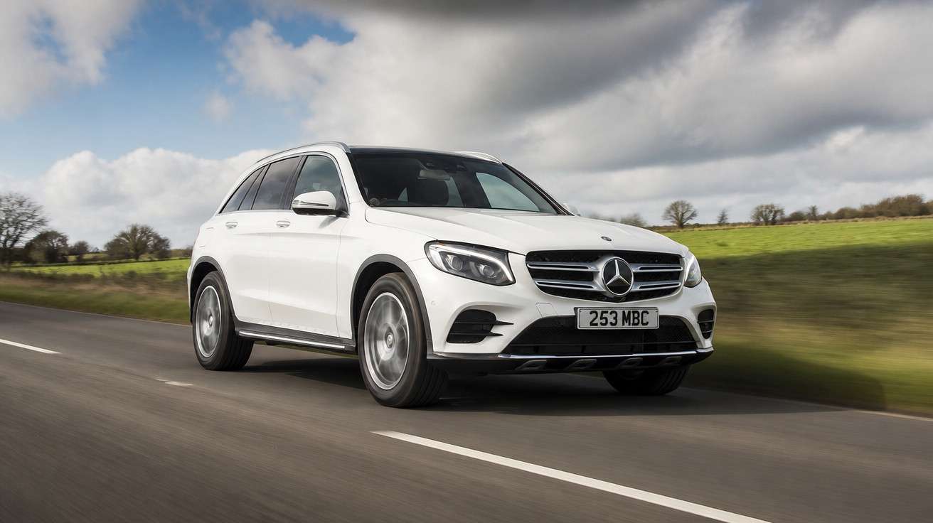 Carro SUV branco Mercedes-Benz em estrada asfaltada com céu nublado e campo verde ao fundo.