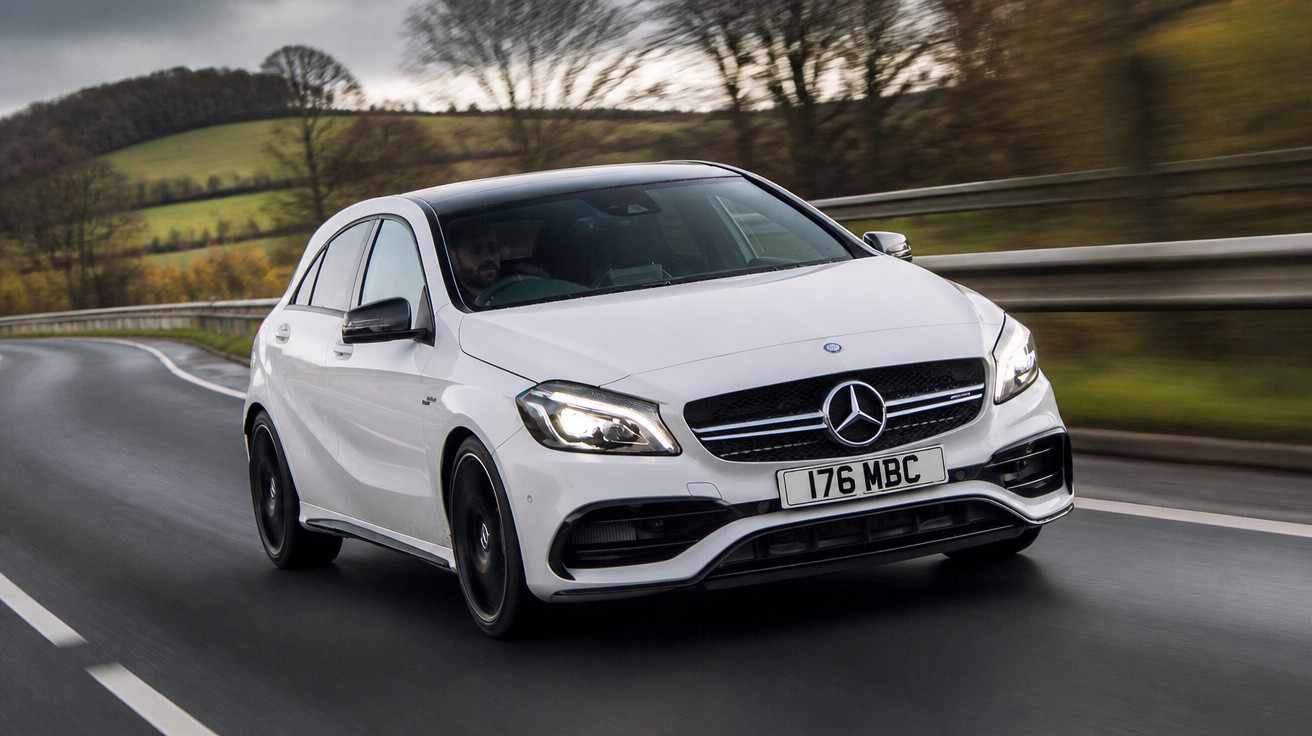 Carro Mercedes-Benz branco em movimento em estrada sinuosa com árvores e colinas ao fundo.