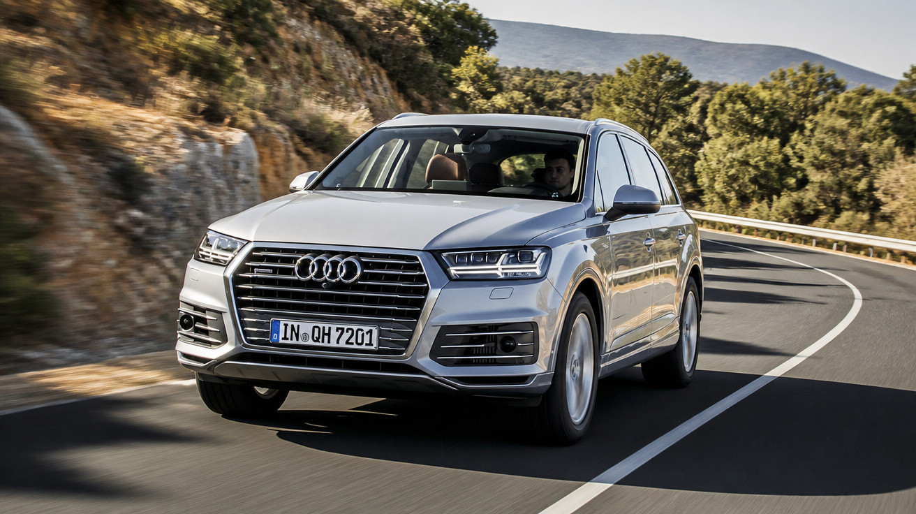 Audi Q7 prata em curva de estrada com motorista, paisagem com árvores e montanhas ao fundo.