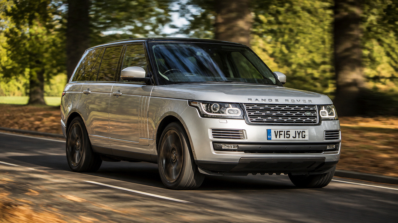 SUV prata Range Rover em estrada cercada por árvores em área ensolarada e com folhas secas no chão.