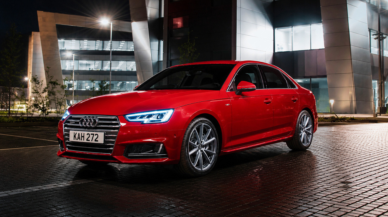 Carro vermelho Audi estacionado em área urbana iluminada à noite com prédio moderno ao fundo.