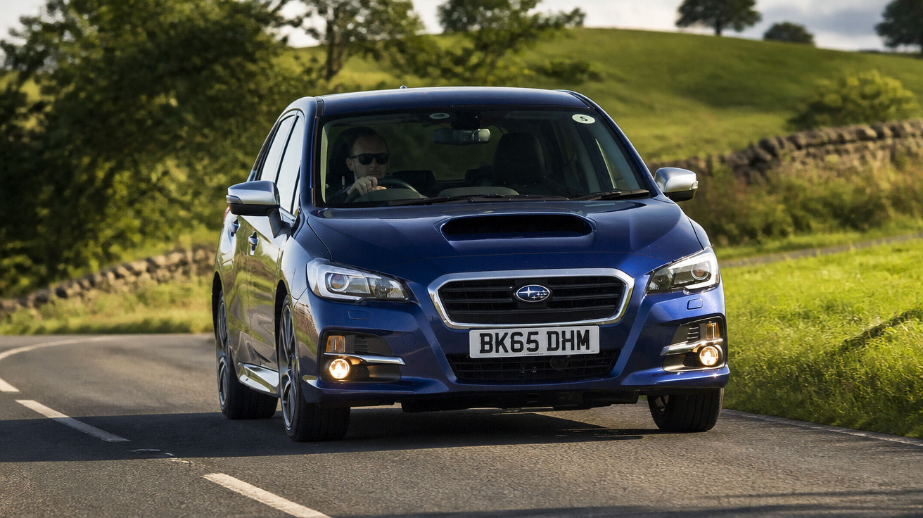 Carro azul Subaru com faróis ligadas dirigindo em estrada rural cercada de árvores e gramado.
