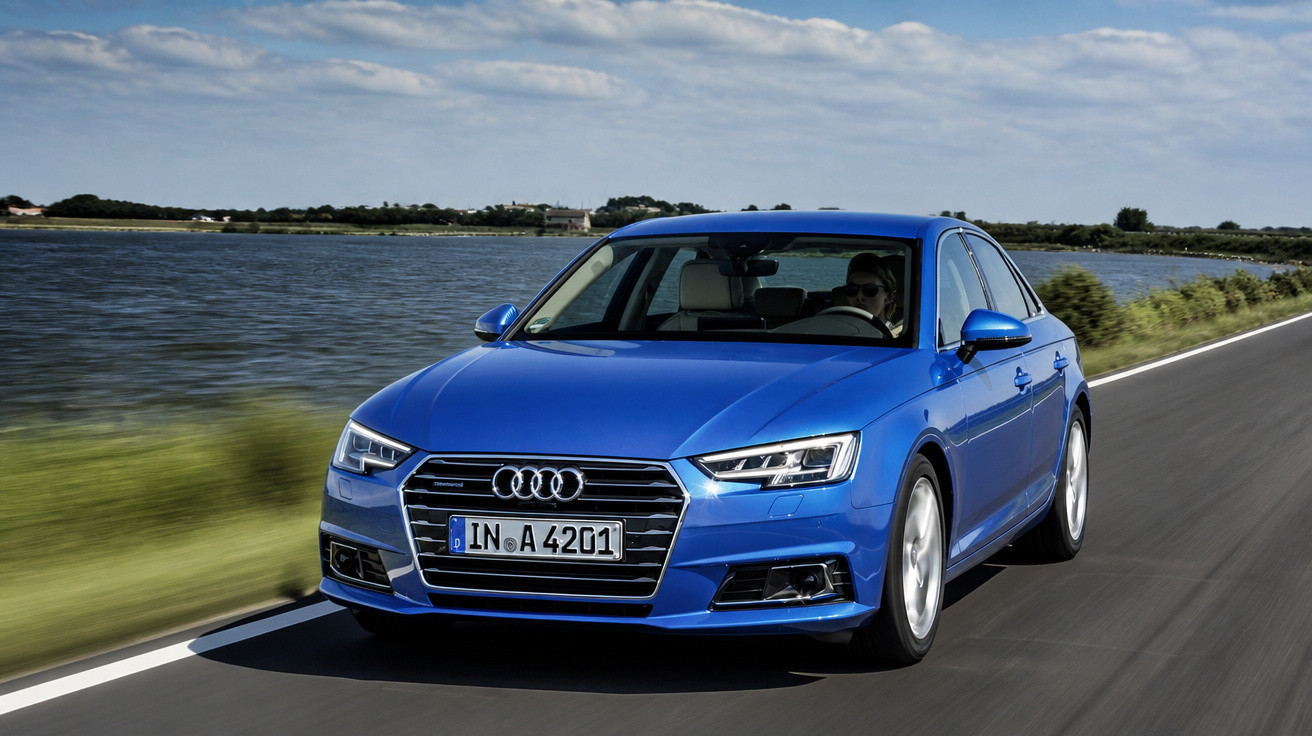 Carro azul Audi A4 circulando em estrada próxima a corpo d'água sob céu com nuvens.