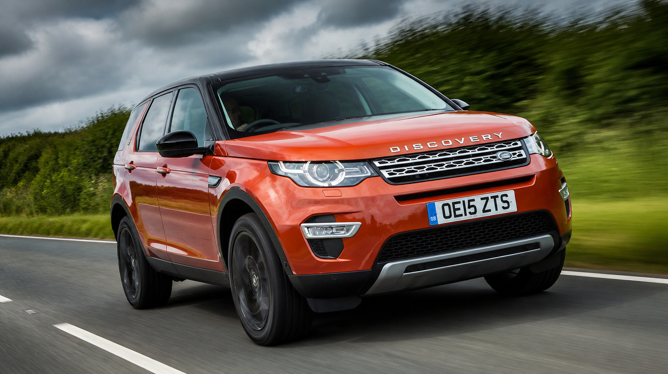 SUV Land Rover Discovery Sport laranja em estrada com céu nublado e vegetação ao fundo.