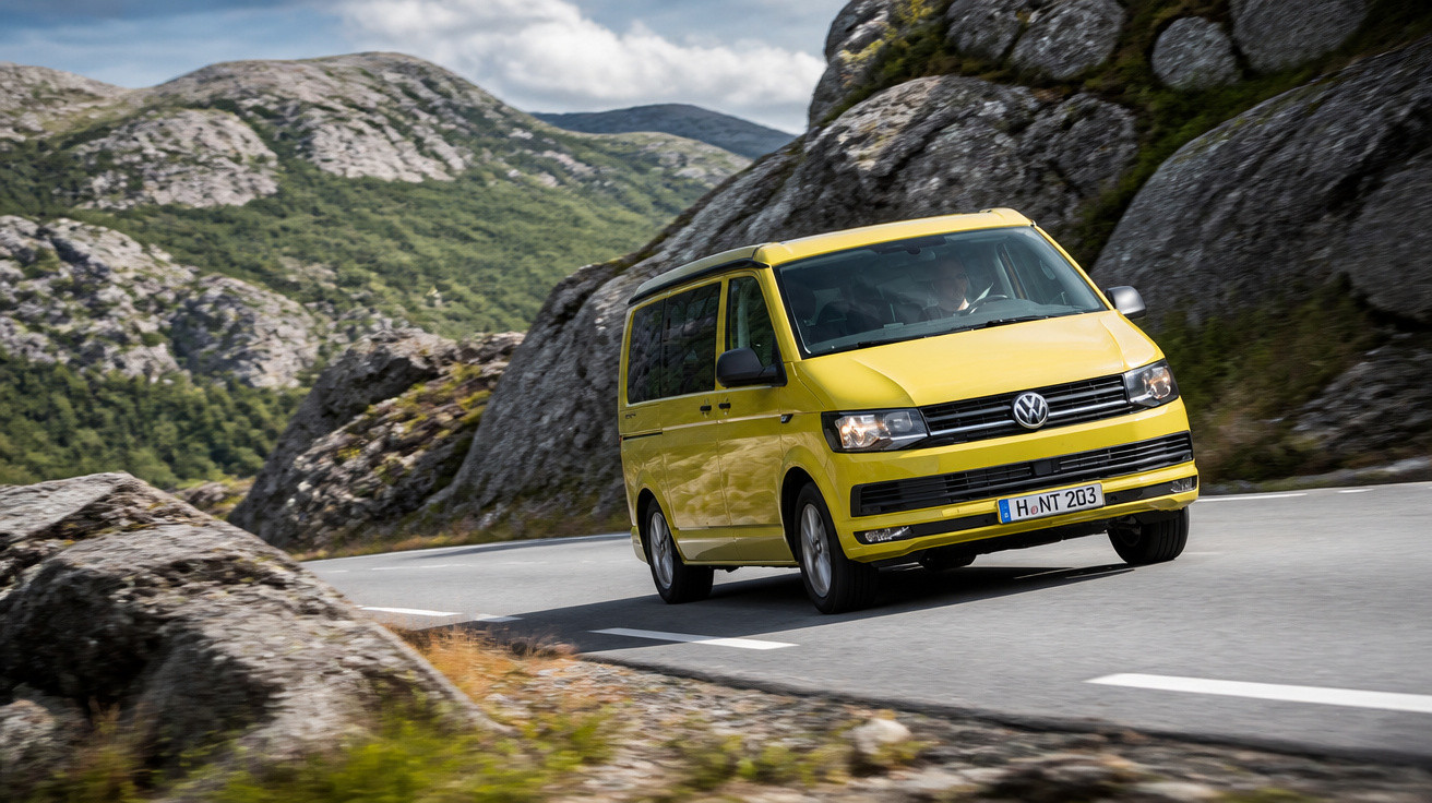 Van amarela Volkswagen em estrada sinuosa cercada por montanhas rochosas e vegetação verde.