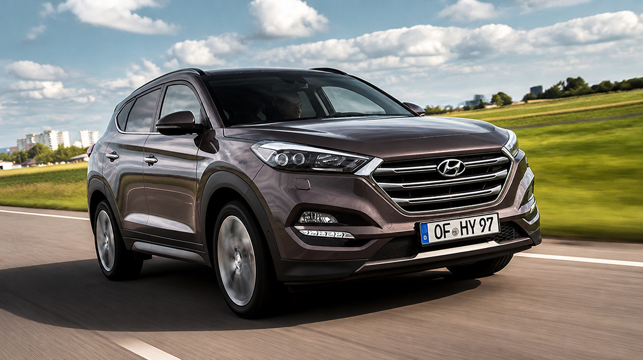 SUV Hyundai Tucson cinza escuro em estrada asfaltada com campo verde ao fundo em dia ensolarado.