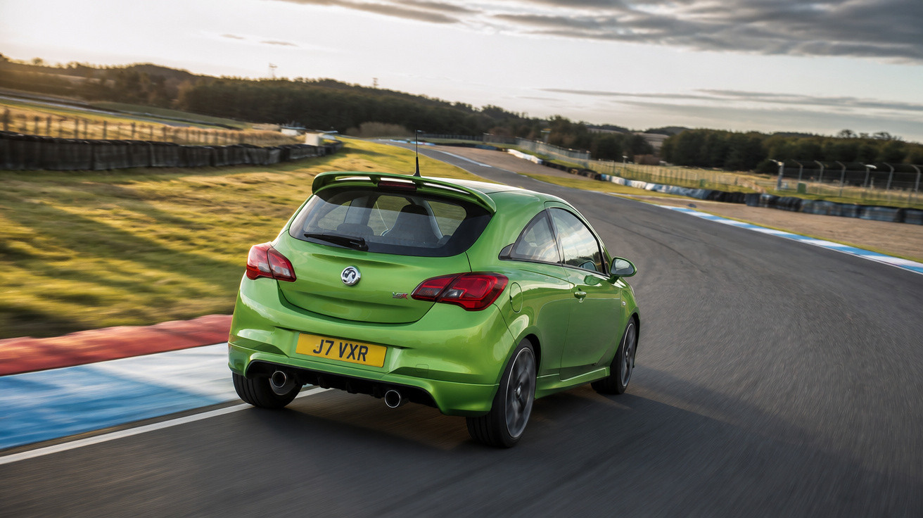 Carro esportivo verde em pista de corrida asfaltada com áreas gramadas e céu nublado ao fundo.