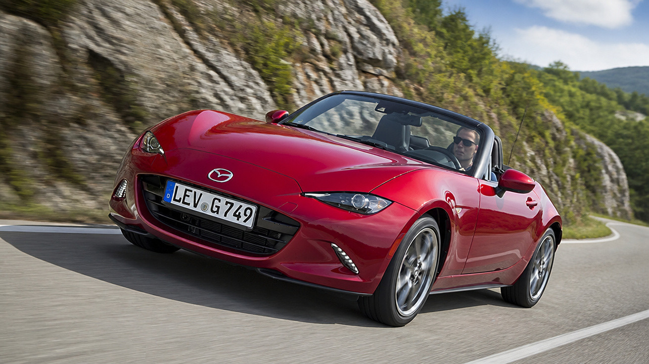 Carro conversível esportivo vermelho Mazda MX-5 dirigindo em estrada sinuosa com montanhas ao fundo.