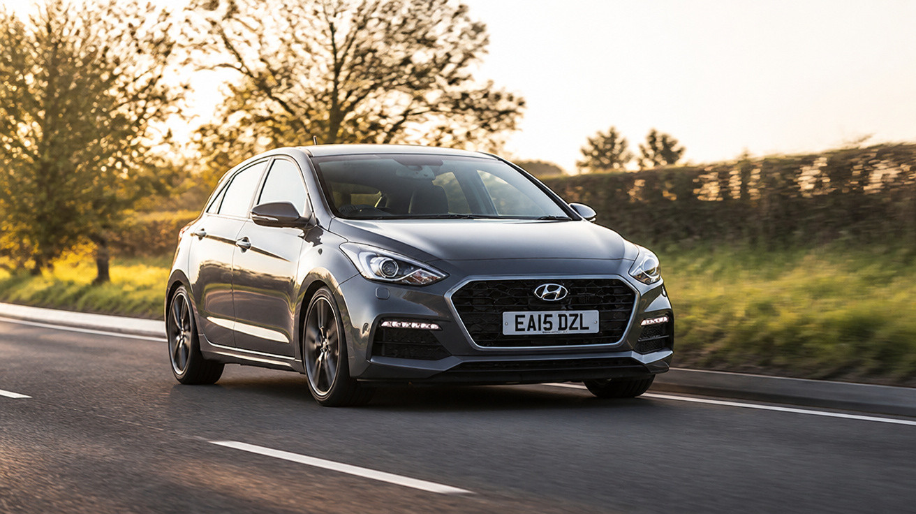 Carro hatchback cinza da Hyundai trafegando em estrada asfaltada ao entardecer com árvores ao fundo.