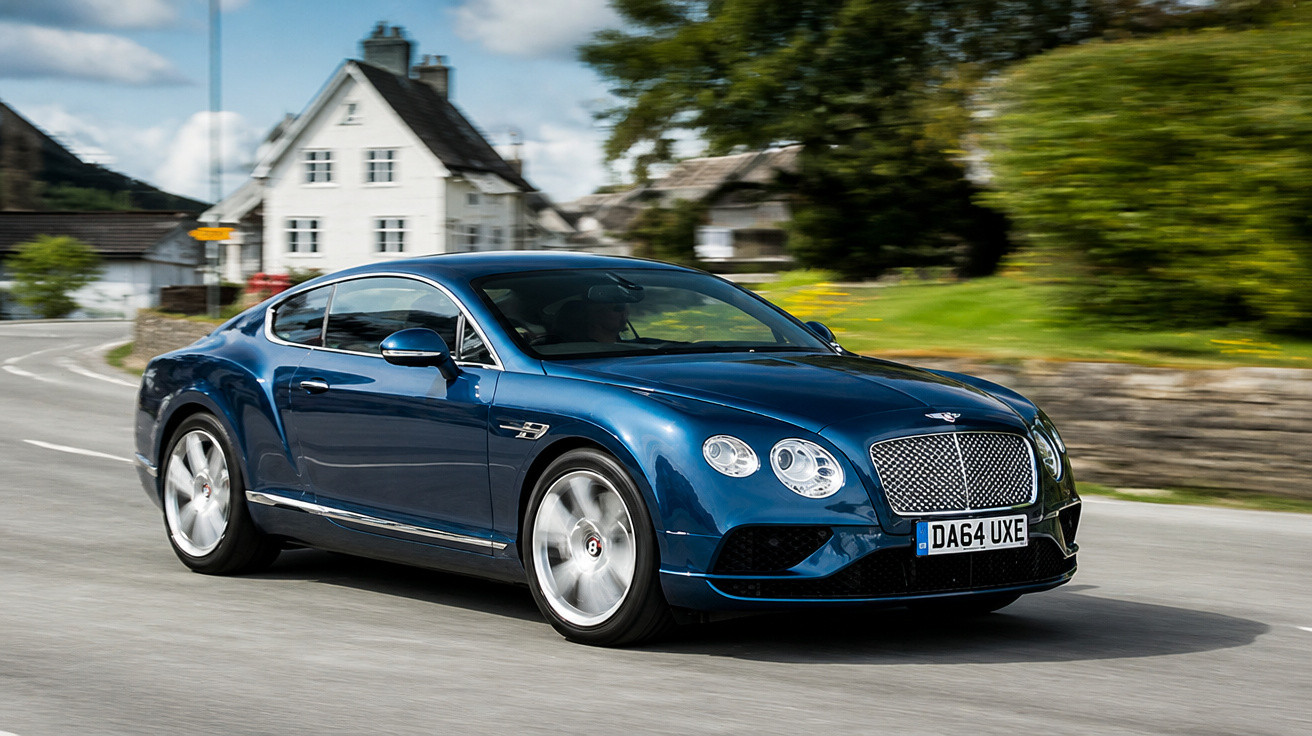 Carro esportivo azul escuro em alta velocidade numa estrada com casas e vegetação ao fundo.