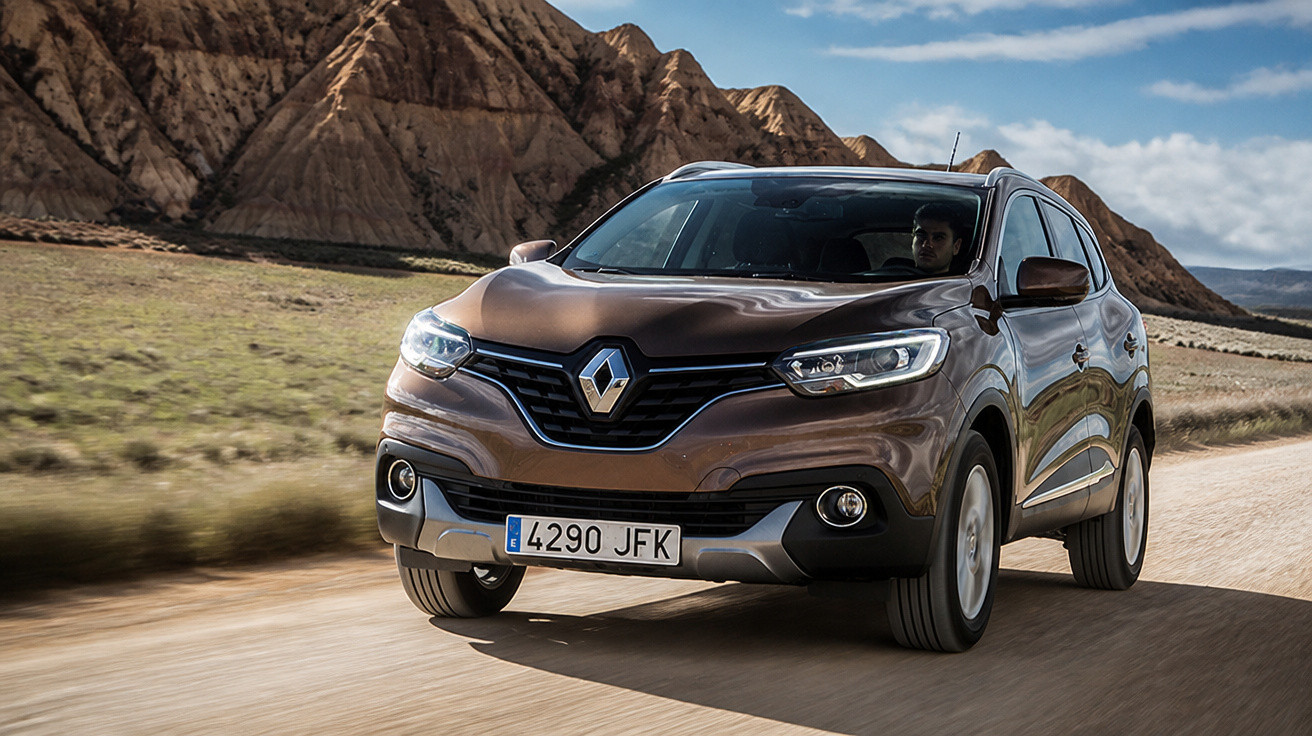 Carro SUV Renault marrom dirigindo em estrada de terra com montanhas ao fundo sob céu azul.