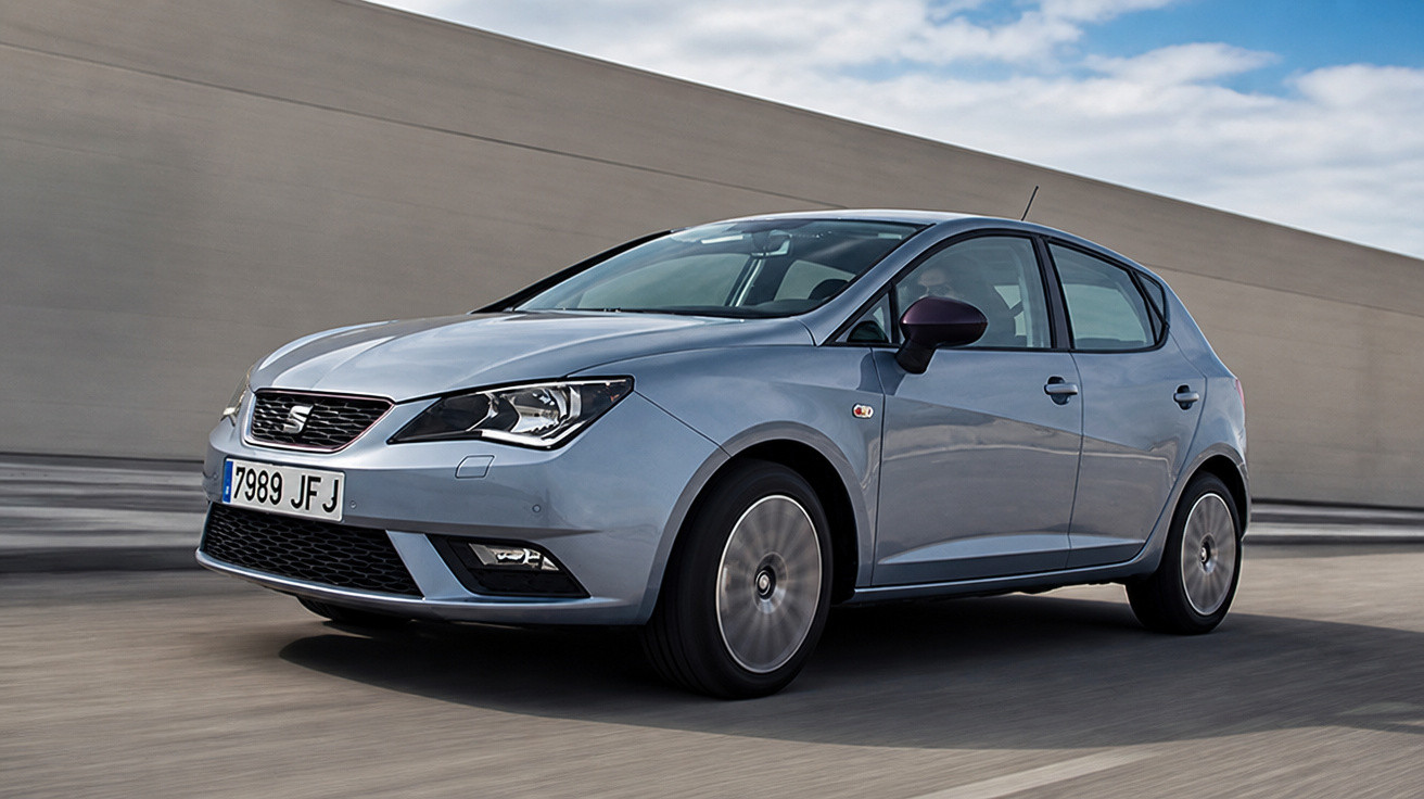 Carro hatchback cinza em movimento em uma estrada com muro cinza ao fundo.