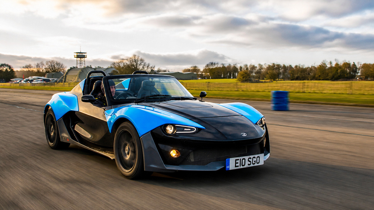 Carro esporte conversível azul e preto em movimento em pista de corrida sob céu nublado.