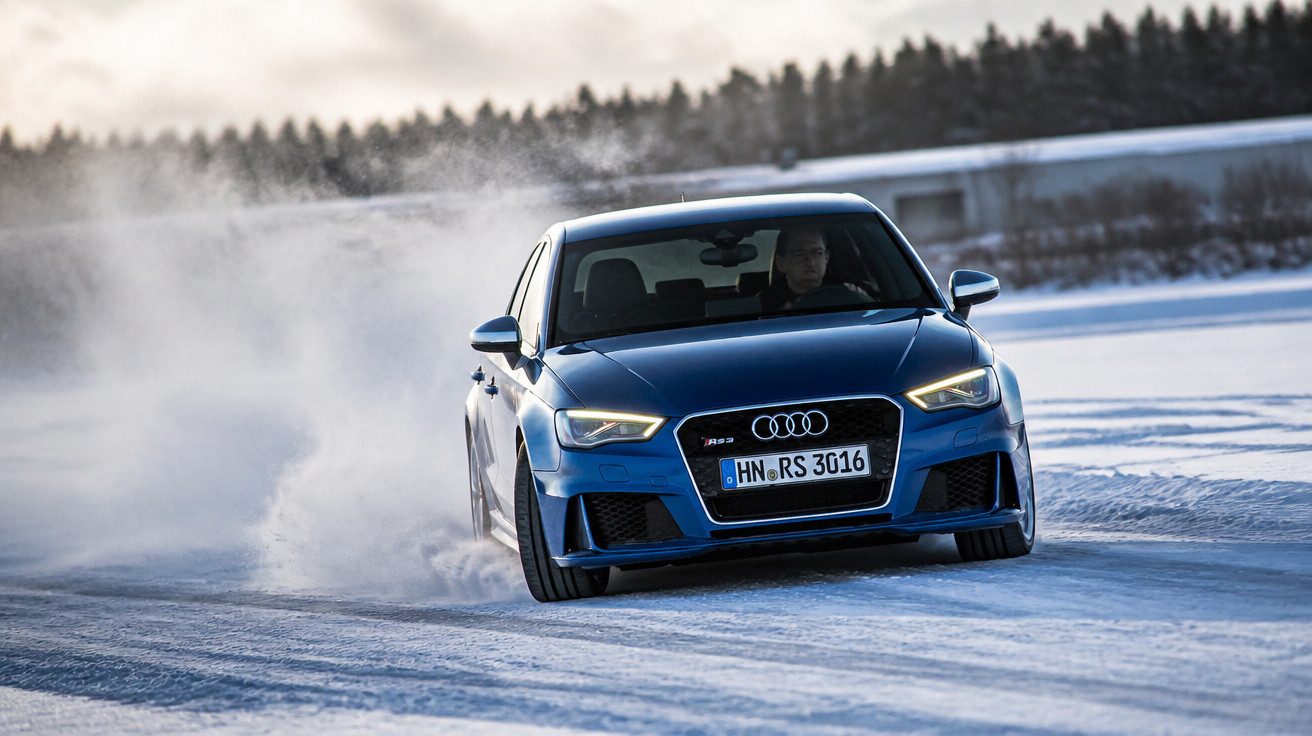 Carro azul Audi em alta velocidade derrapando na neve em estrada gelada com árvores ao fundo.