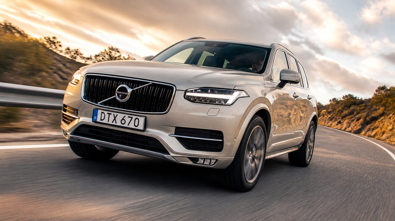 Carro Volvo SUV prata dirigindo em estrada sinuosa com árvores e céu nublado ao fundo.