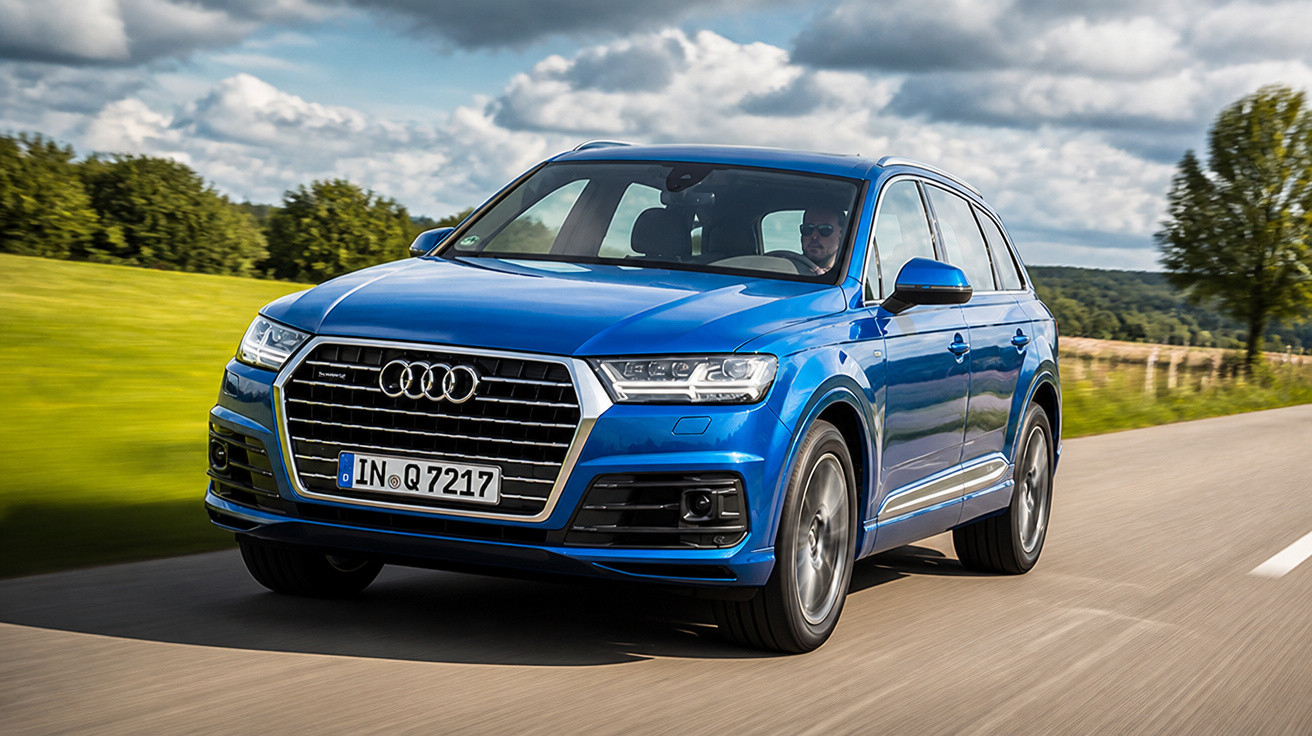 Carro SUV azul Audi Q7 circulando em estrada com campo verde e céu nublado ao fundo