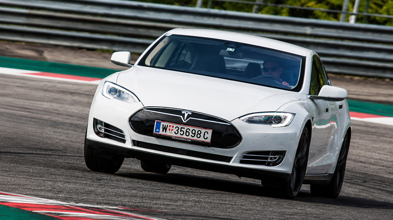 Carro elétrico Tesla branco fazendo curva em pista de corrida com barreira de proteção metálica.