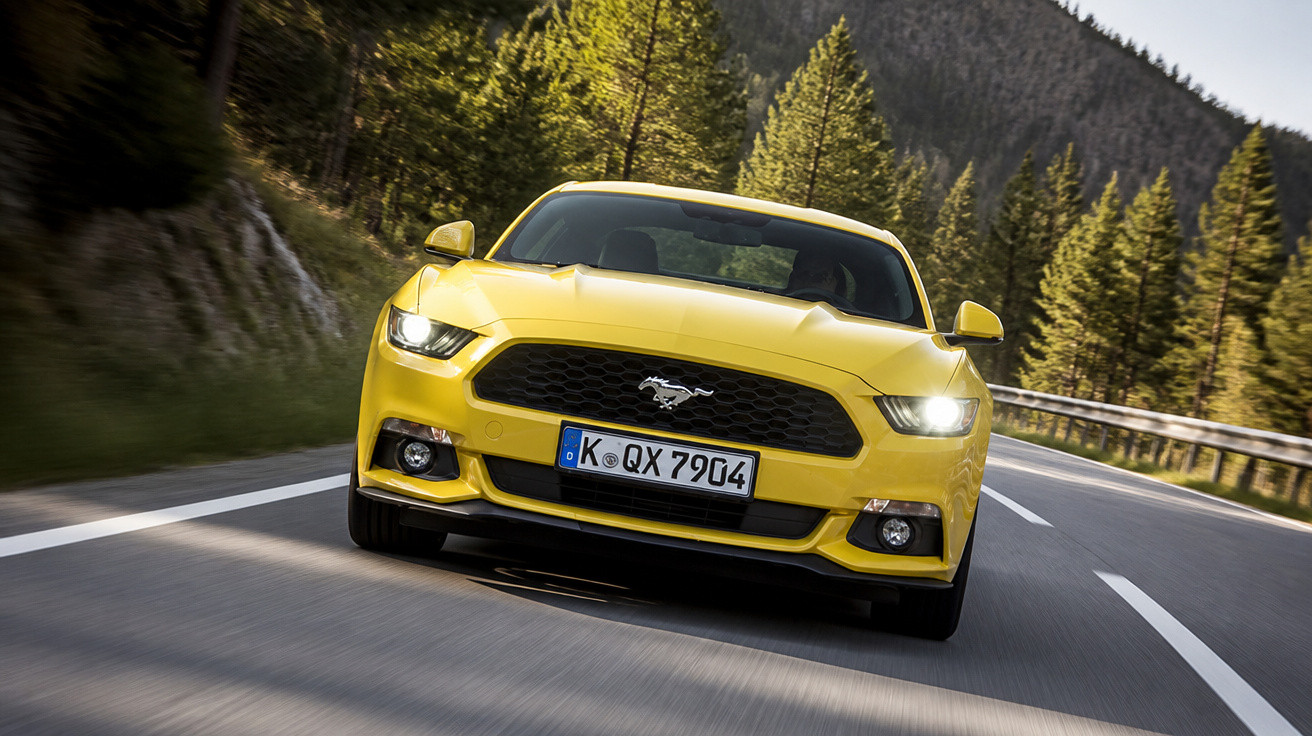 Carro esportivo amarelo Ford Mustang em estrada rodeada por árvores e montanhas.