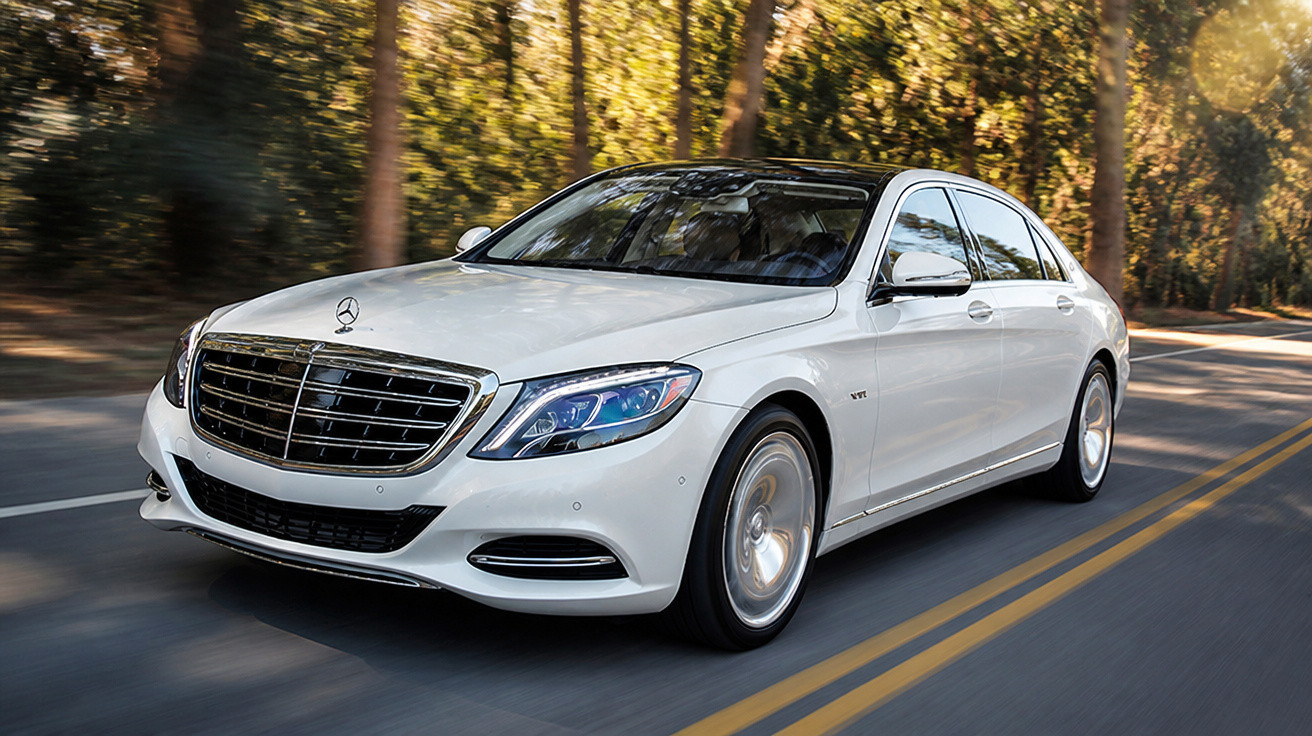 Carro sedã branco da Mercedes-Benz em movimento em estrada cercada por árvores ao entardecer.